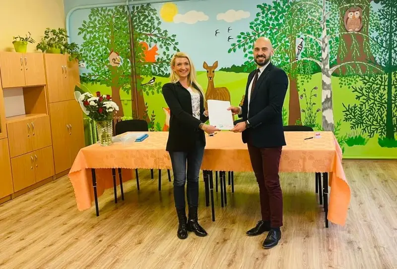 Zdjęcie w orientacji poziomej przedstawia dwie osoby stojące wewnątrz przedszkola, trzymające wspólnie biały dokument. Kobieta po lewej stronie ubrana jest w czarną marynarkę, jeansy i ciemne botki, natomiast mężczyzna po prawej ma na sobie elegancki granatowy garnitur z bordowym krawatem i ciemne spodnie. Za nimi znajduje się stół przykryty pomarańczowym obrusem, na którym stoi wazon z bukietem czerwono-białych kwiatów. Tło stanowi kolorowy mural przedstawiający leśny krajobraz z drzewami, sarną, sową i innymi elementami przyrody, namalowany na ścianie sali. Po lewej stronie kadru widoczne są drewniane szafki w jasnym odcieniu, które dopełniają wystrój wnętrza placówki edukacyjnej.
