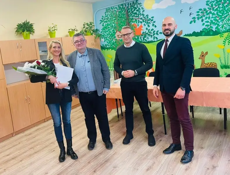 Zdjęcie o orientacji poziomej przedstawia cztery uśmiechnięte osoby stojące w sali przedszkolnej, ustawione w jednym rzędzie na tle malowidła ściennego z motywem leśnym. Po lewej stronie stoi kobieta w ciemnej marynarce i jeansach, trzymająca bukiet kwiatów oraz białą teczkę, a obok niej znajdują się trzej mężczyźni w eleganckich strojach. Tło tworzą jasne szafki z kwiatami doniczkowymi w górnej części oraz kolorowy mural przedstawiający drzewa, sarenki i ptaki nad stołem przykrytym brzoskwiniowym obrusem. Kompozycja zdjęcia jest symetryczna, a postacie zajmują większą część kadru na jasnej, drewnianej podłodze. Oświetlenie jest równomierne, co pozwala na wyraźne wyeksponowanie bohaterów oraz przyjaznego charakteru wnętrza placówki edukacyjnej.