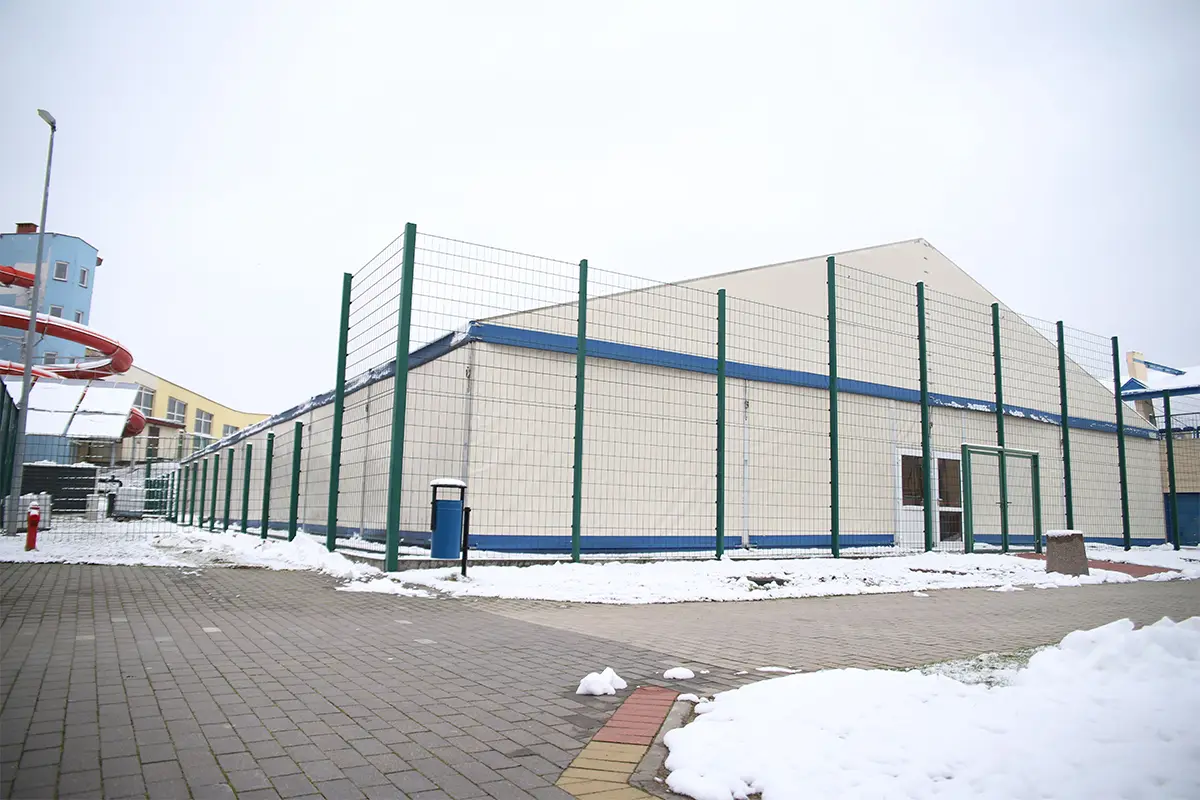 Zdjęcie przedstawia dużą halę sportową o konstrukcji namiotowej w formacie poziomym, otoczoną wysokim ogrodzeniem z siatki. Obiekt posiada jasne, kremowe ściany z wyraźnymi niebieskimi pasami wykończeniowymi i znajduje się na terenie wyłożonym kostką brukową przykrytą miejscami śniegiem. W lewej części kadru widoczna jest charakterystyczna czerwona zjeżdżalnia wodna należąca do pobliskiego aquaparku oraz wysoka latarnia. Na pierwszym planie po lewej stronie umieszczony jest niebieski kosz na śmieci, a po prawej stronie znajduje się betonowy słup przy wejściu do hali. Całość kompozycji charakteryzuje się dość szerokim kątem widzenia, obejmującym cały boczny profil budynku oraz otaczającą go zimową infrastrukturę.