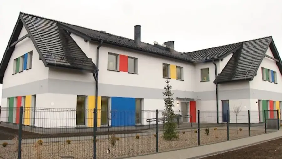 Zdjęcie przedstawia nowoczesny budynek użyteczności publicznej lub przedszkole w układzie poziomym o proporcjach boków zbliżonych do 16:9. Biała elewacja obiektu jest urozmaicona charakterystycznymi, kolorowymi akcentami w odcieniach czerwieni, niebieskiego, zielonego i żółtego, które znajdują się między oknami. Budynek posiada dwuspadowy dach pokryty ciemną dachówką oraz szary pas dekoracyjny biegnący wzdłuż ścian parteru. Całość otoczona jest metalowym ogrodzeniem panelowym, za którym widać zadbany teren z młodymi nasadzeniami drzew i iglaków. Przed wejściem do budynku widoczna jest rampa dla osób niepełnosprawnych, a przed ogrodzeniem biegnie chodnik z kostki brukowej.